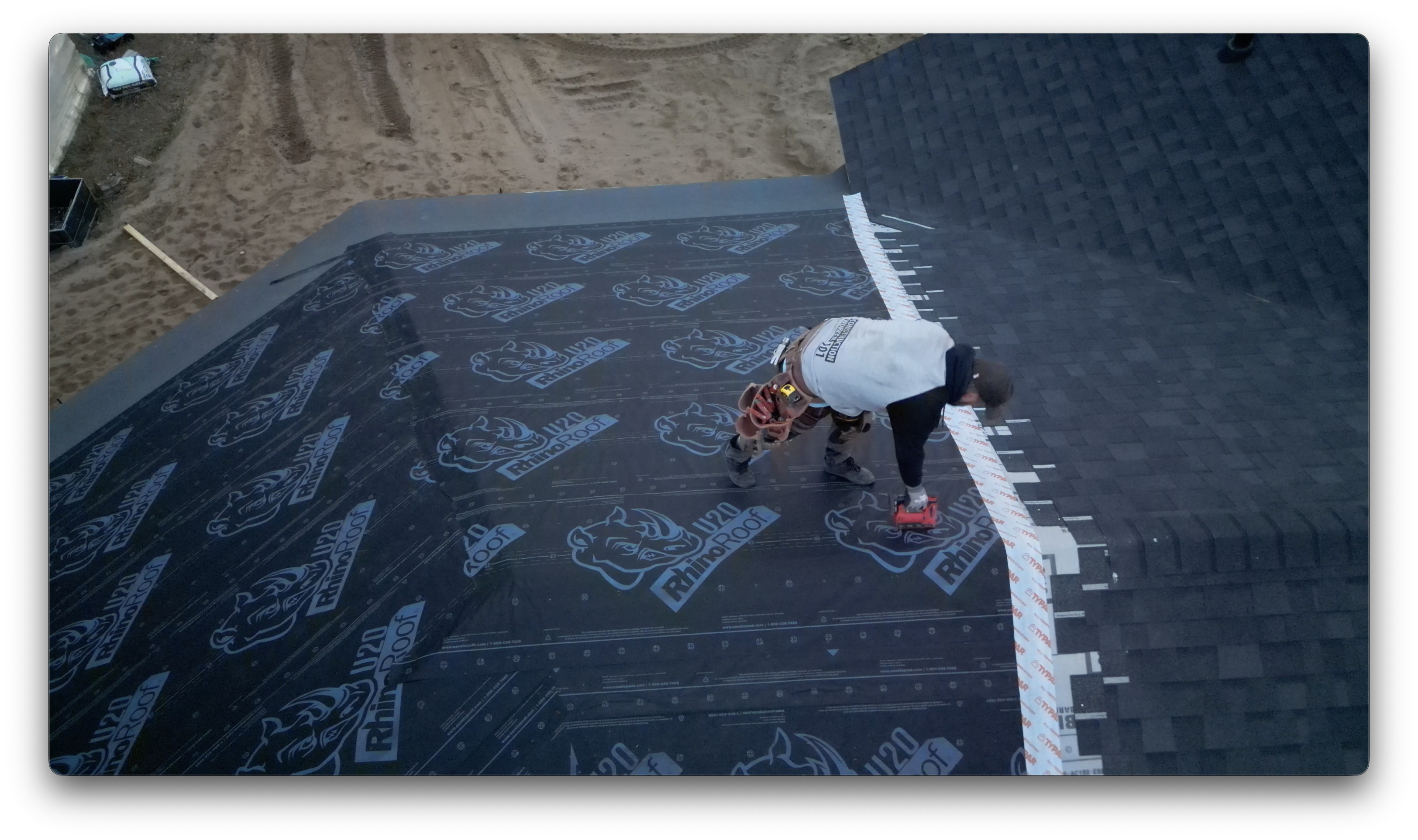 A worker installs shingles along a roof seam while securing underlayment during residential roofing construction.