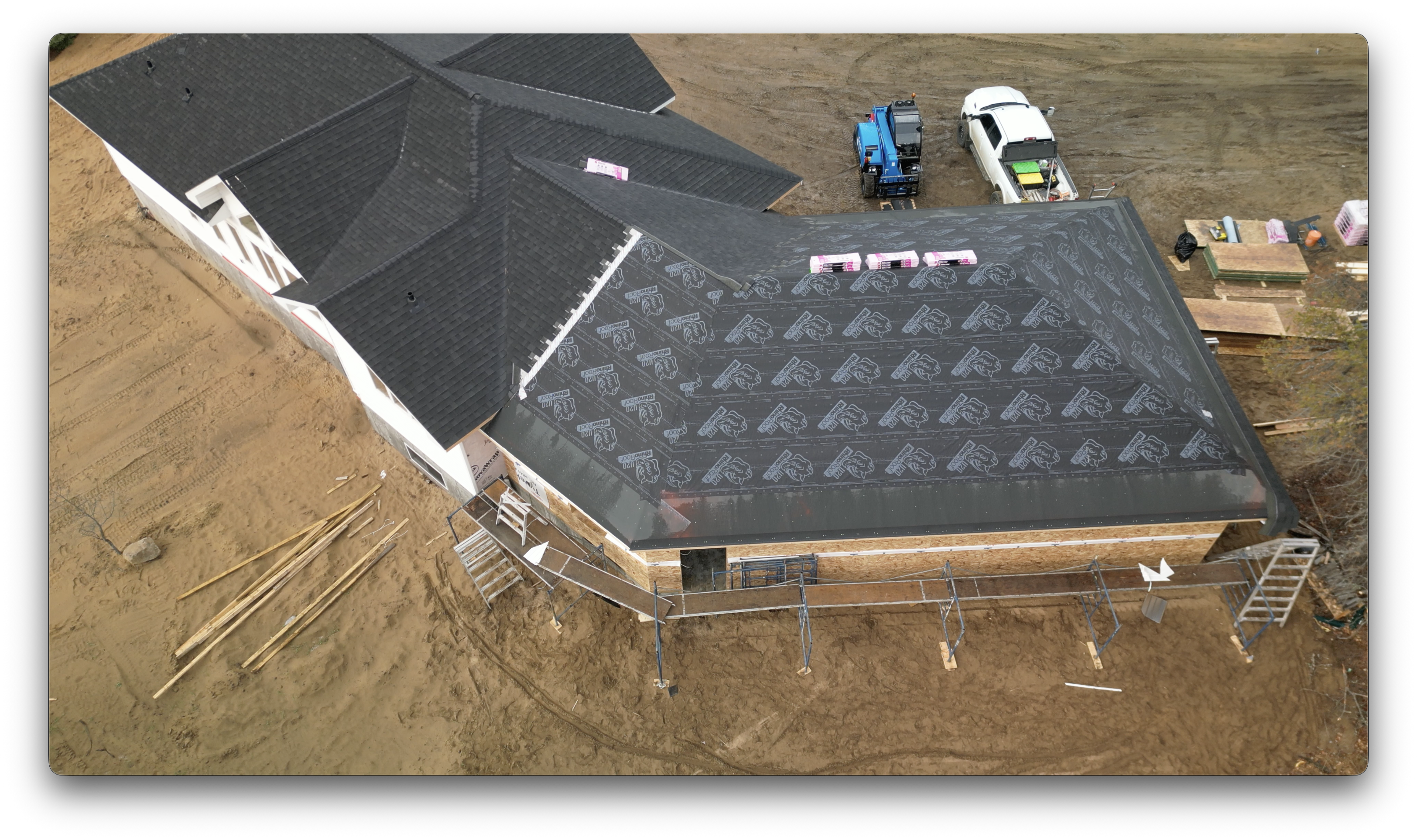 Drone view of a house under construction with shingles installed on part of the roof and roofing underlayment covering the newly built section.