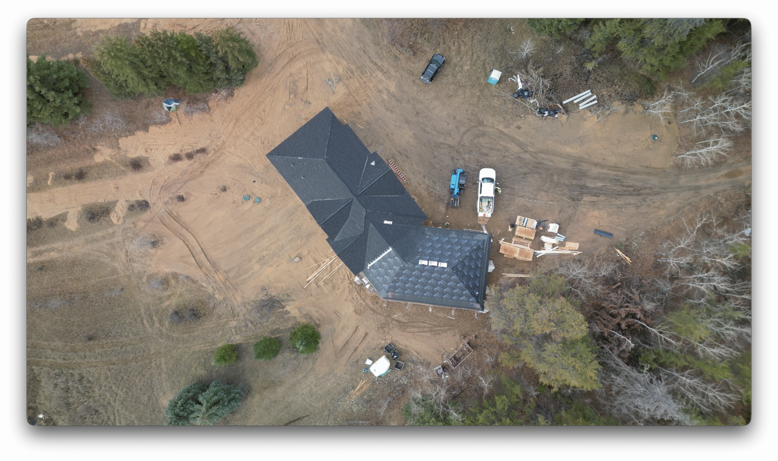 A high drone view of a home under construction, showing newly installed shingles, roofing underlayment, construction vehicles, and building materials spread around the site.