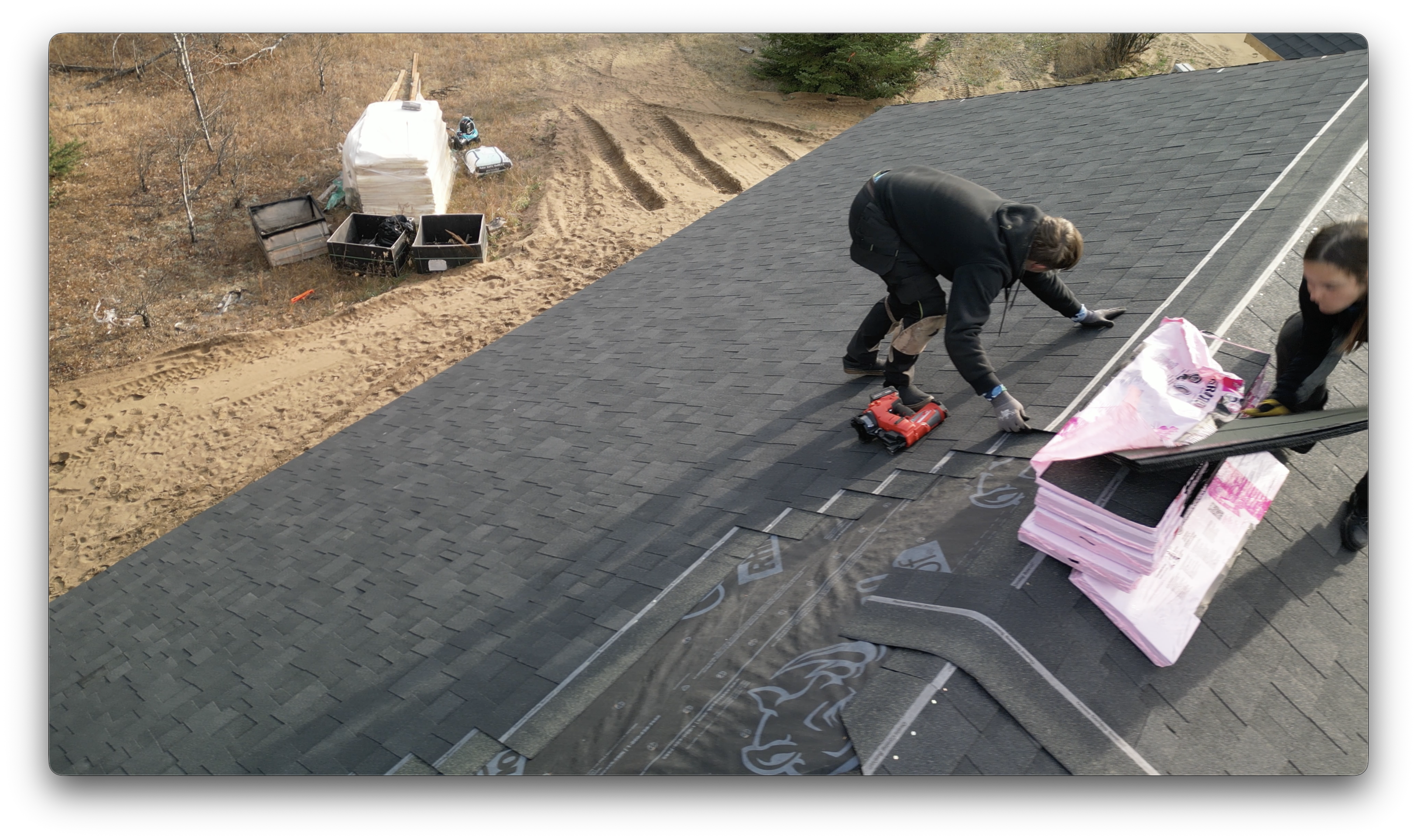 Two roofing workers install shingles on a steep roof, using a nail gun and working beside stacked shingle bundles.