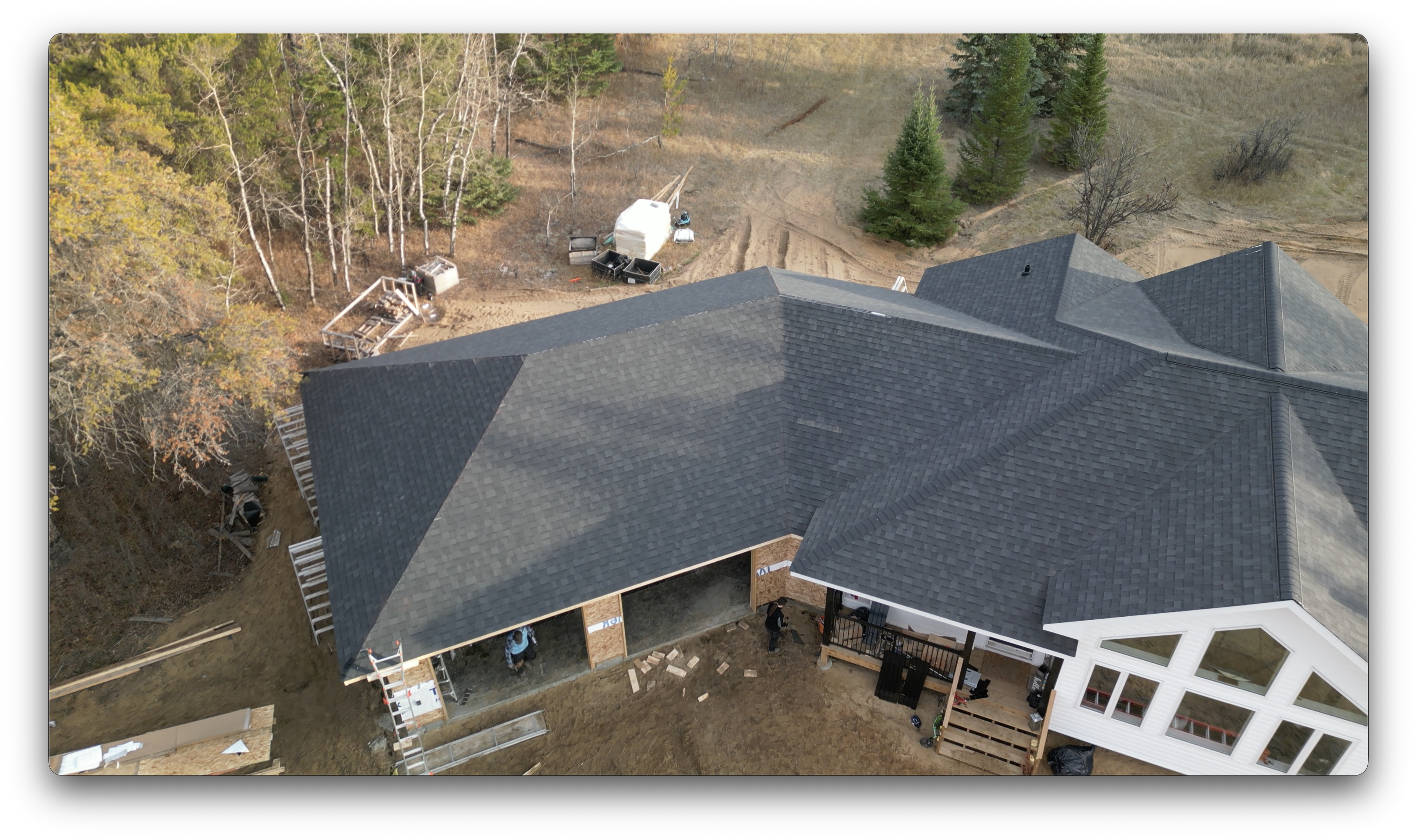 Drone image of a house with newly installed dark asphalt shingles, showing garage openings, workers on-site, and surrounding forested landscape.