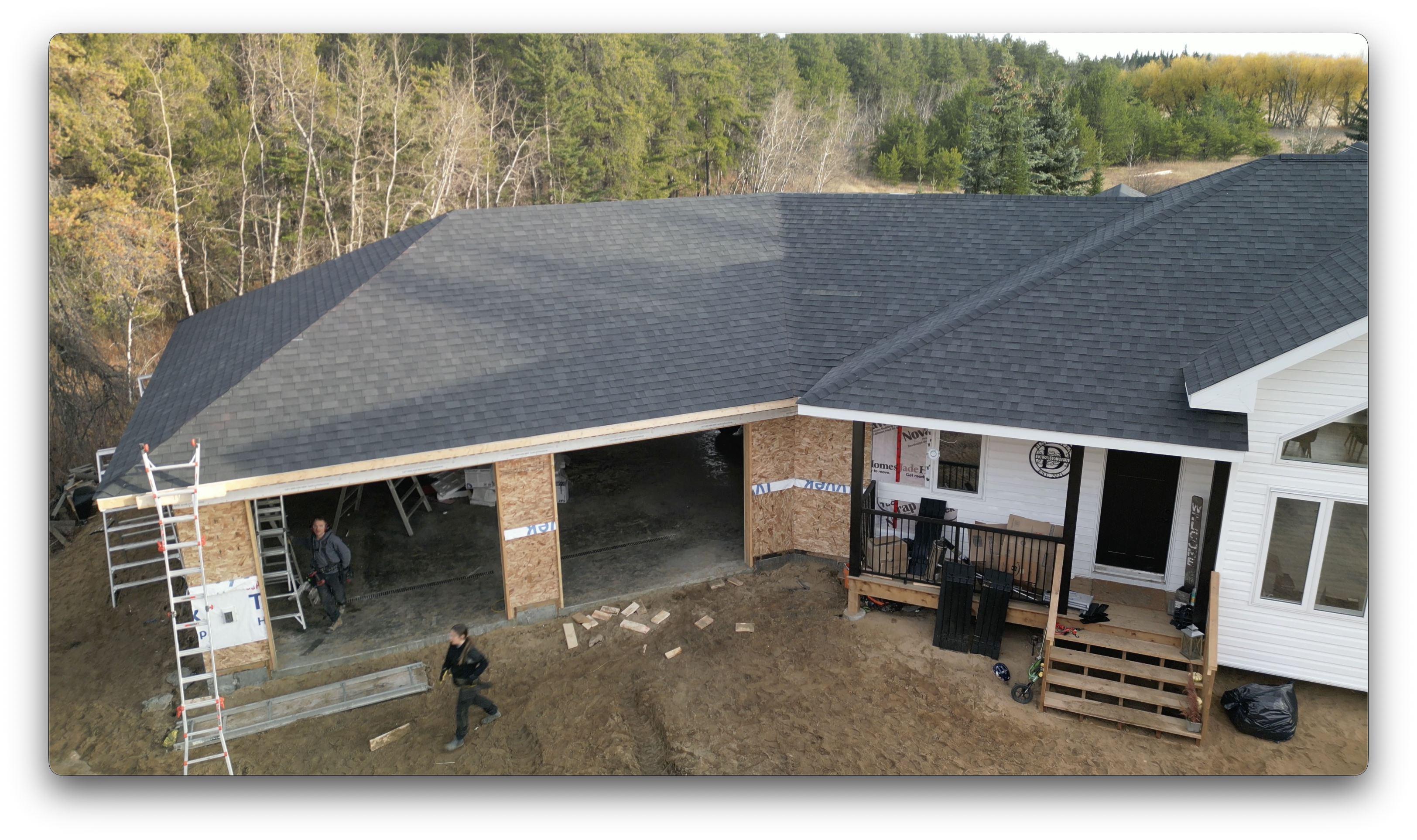 Drone image of a house with newly installed dark asphalt shingles, showing garage openings, workers on-site, and surrounding forested landscape.