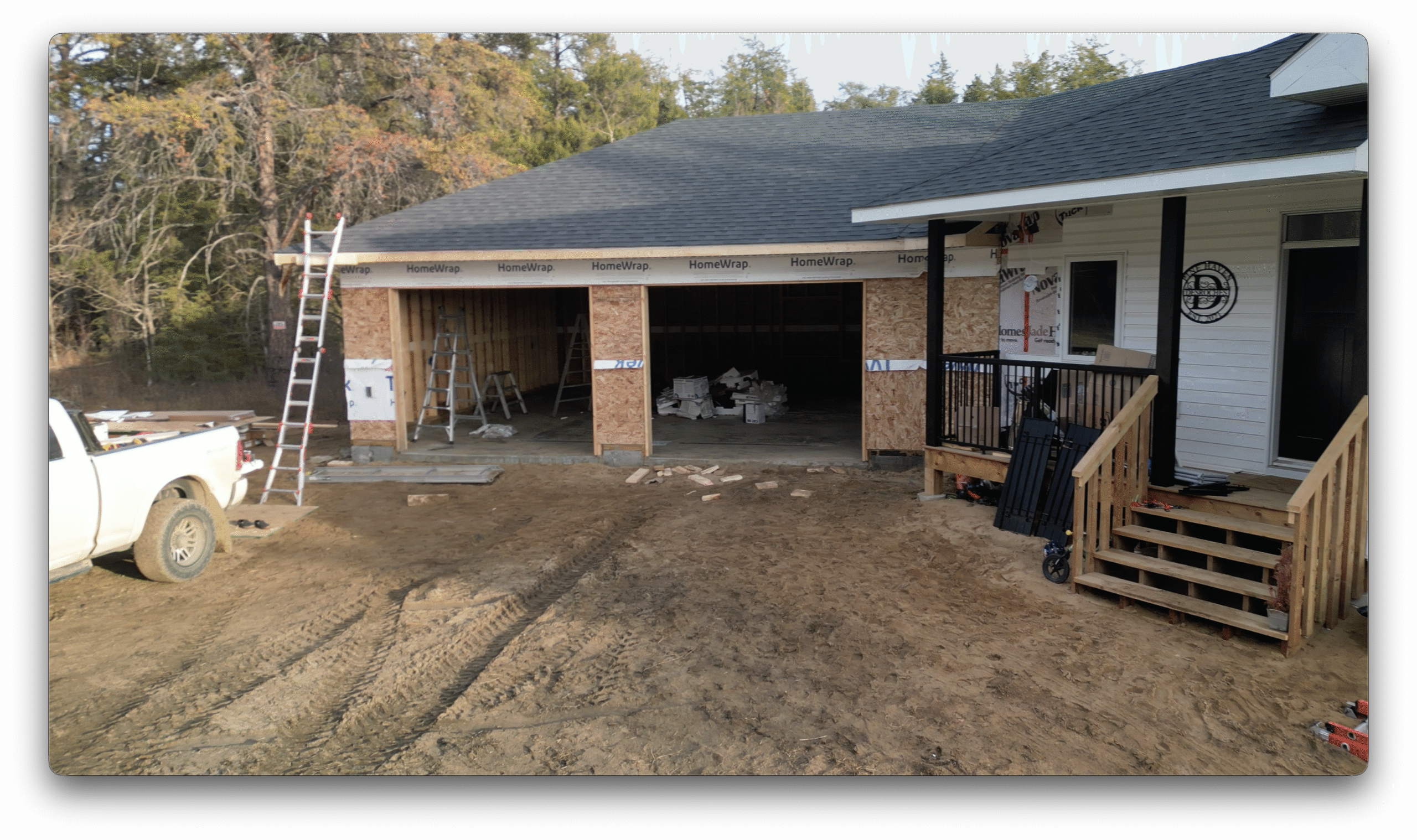 A construction site showing a partially finished garage attached to a house, with ladders, tools, and building materials staged around the entrance.