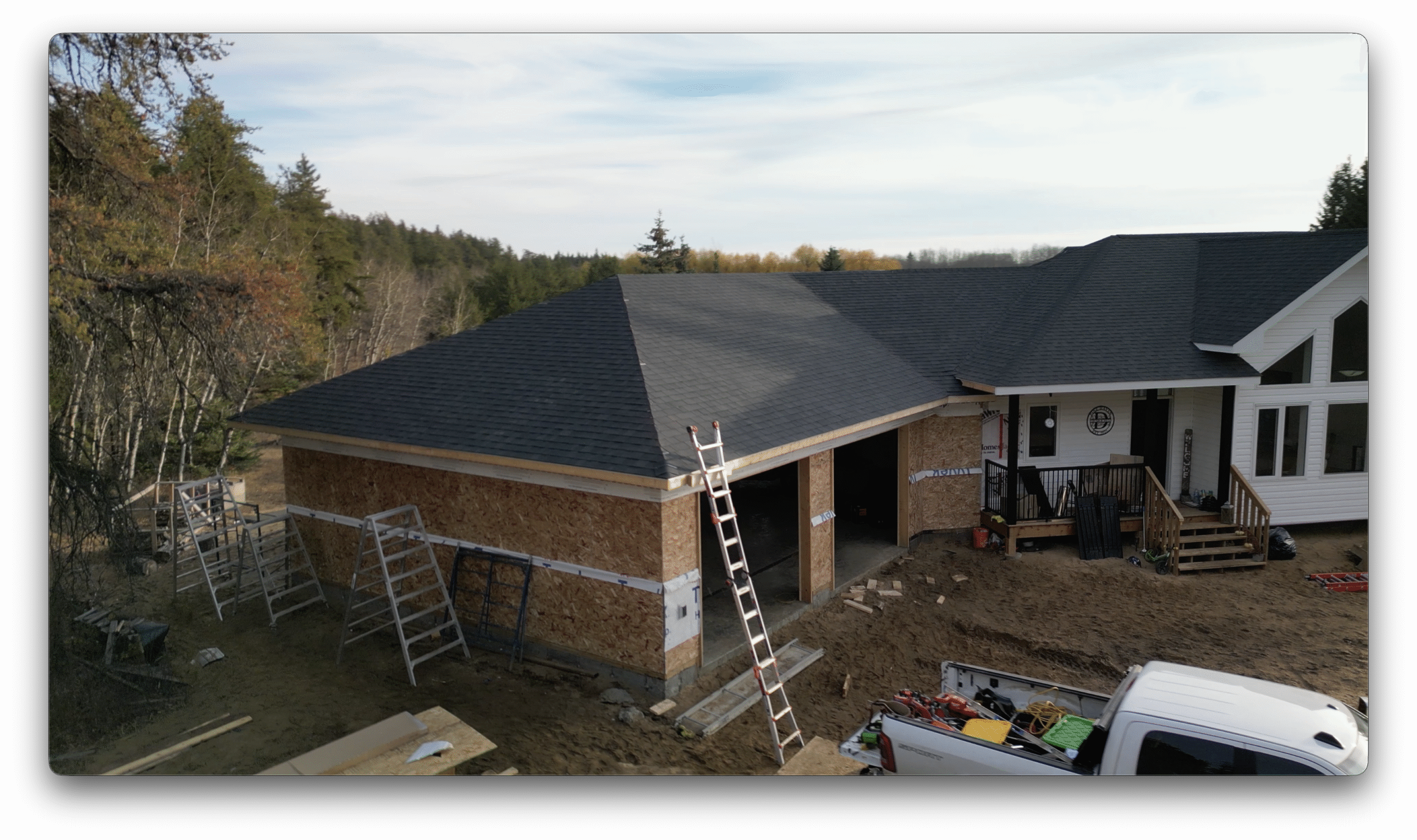 A construction site showing a newly shingled garage roof attached to a house, with ladders, tools, and building materials around the unfinished exterior.