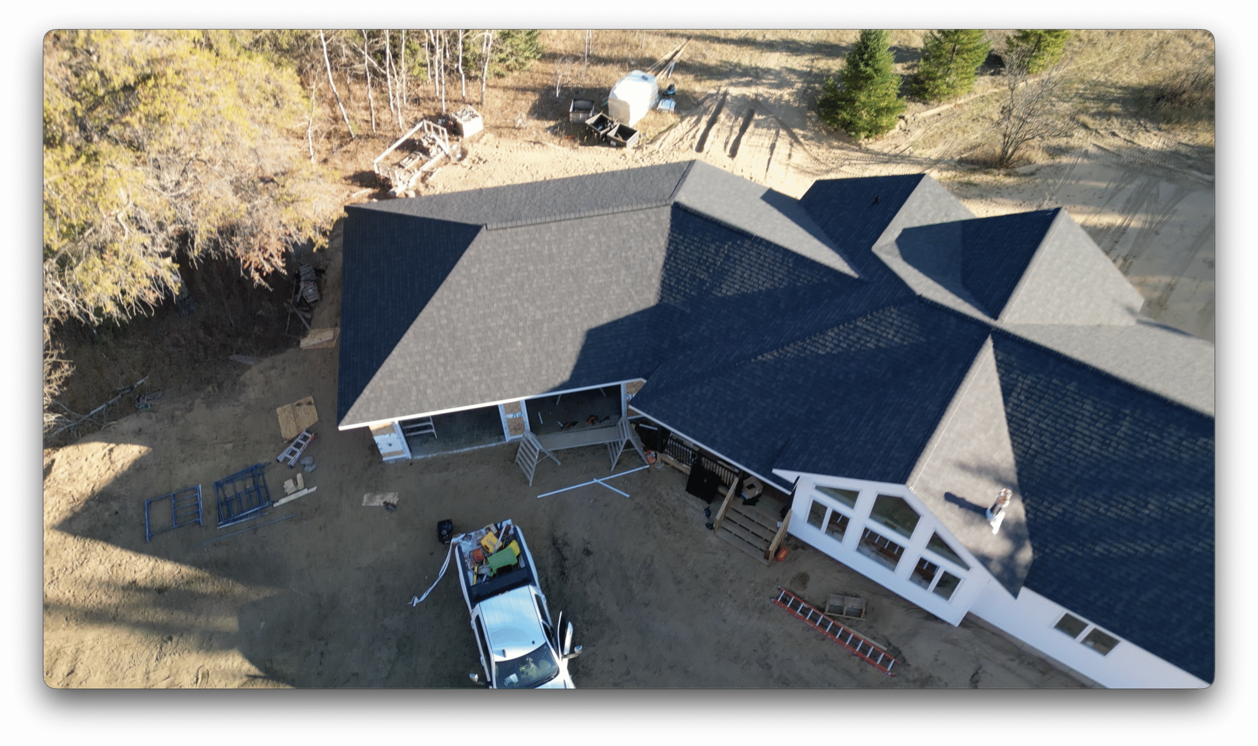 Aerial photo of a completed residential roof showing dark asphalt shingles, a garage structure, and construction equipment on the ground below.