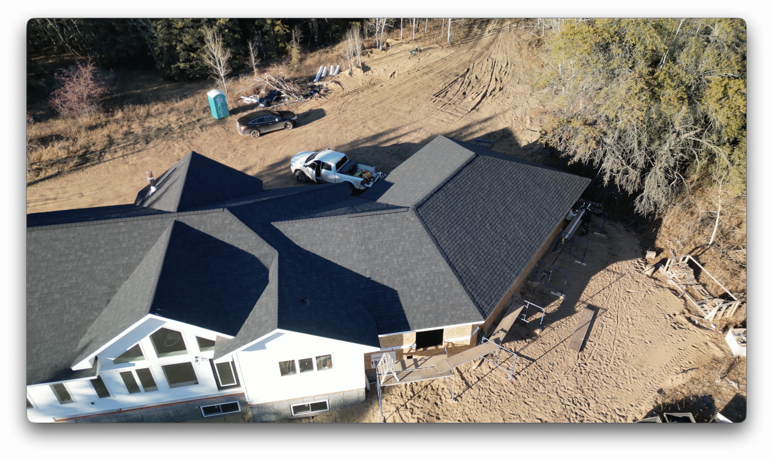 Drone image of a completed shingled roof with surrounding construction materials, vehicles, and natural wooded landscape.