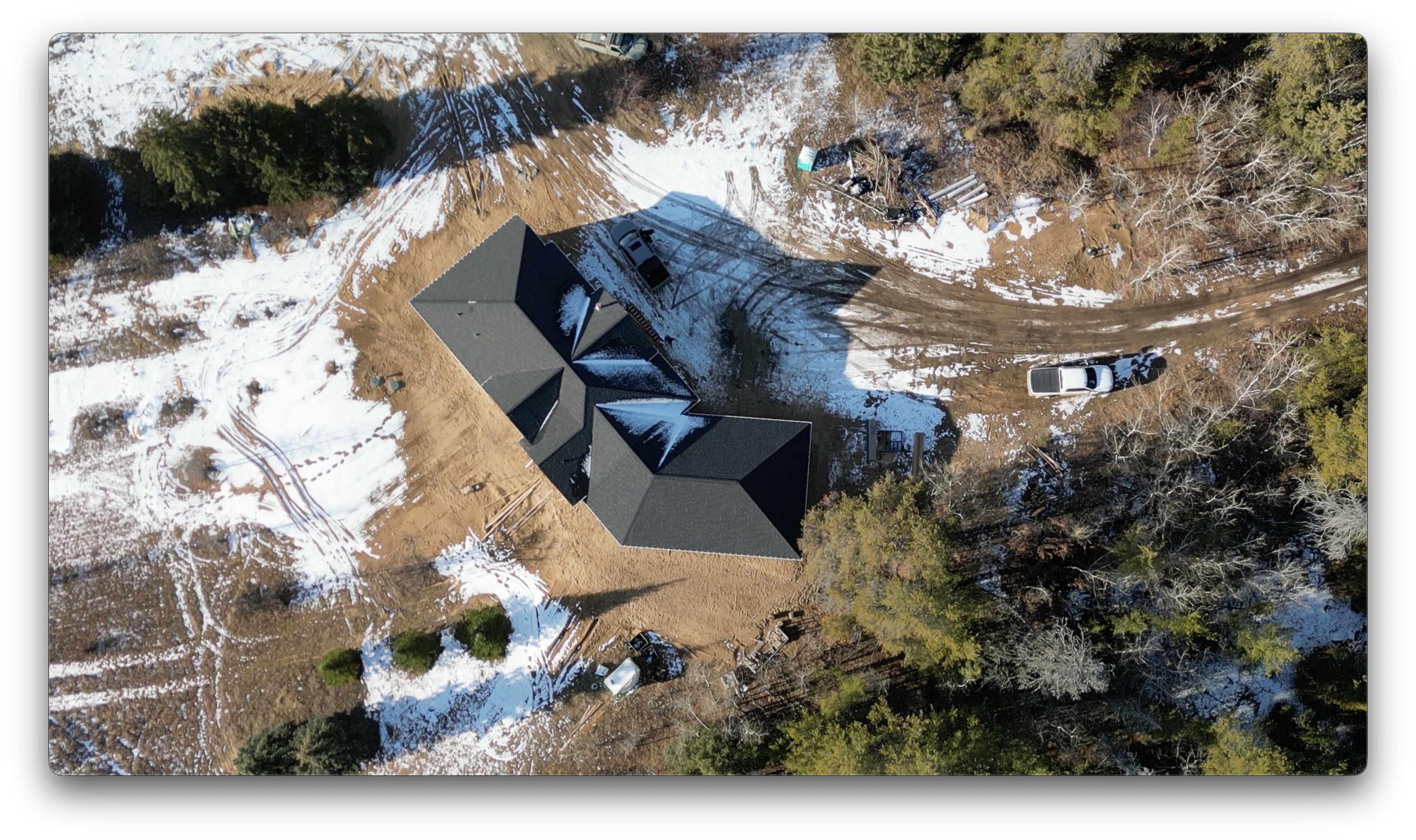 Top-down drone view of a finished roof on a rural residential construction site with light snow on the ground and vehicles parked nearby.