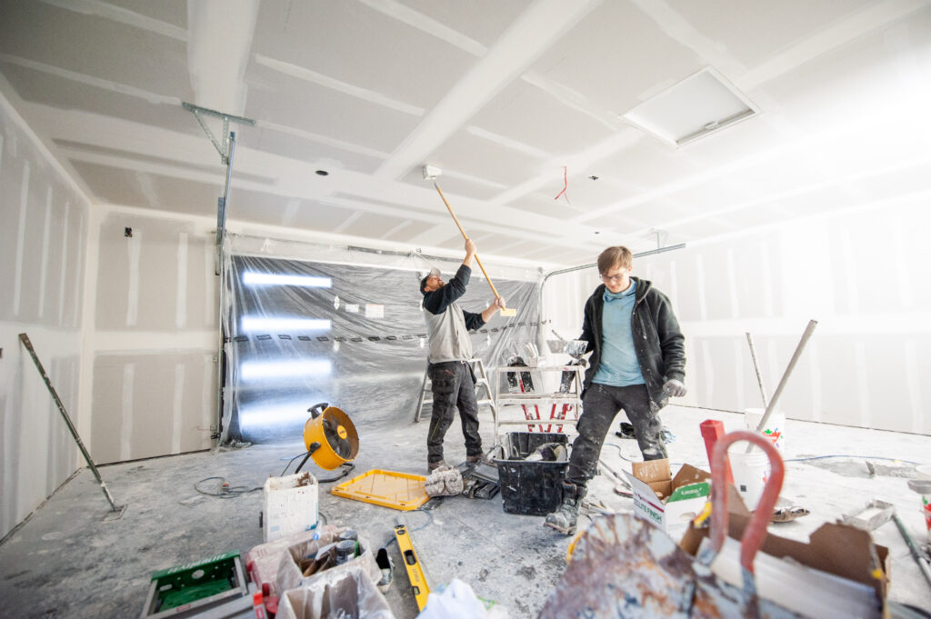 Workers applying drywall mud to walls and ceilings in a finished garage interior.