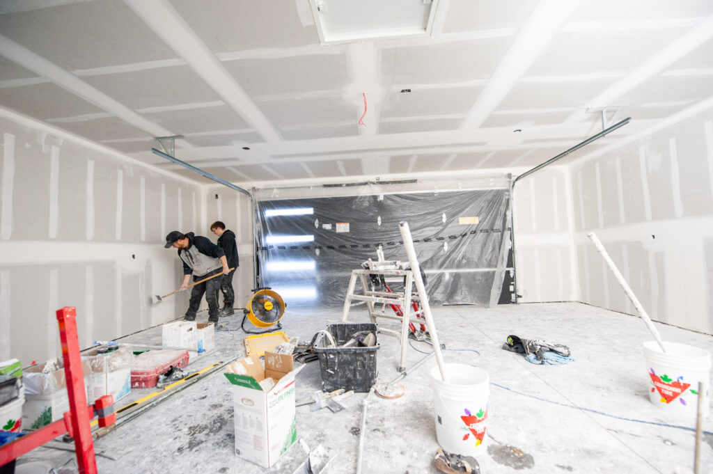 Drywall finishers applying compound to seams inside a fully drywalled two-car garage.