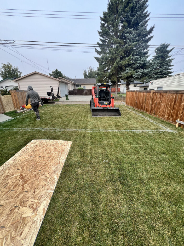 Site preparation for a detached garage build in Edmonton with a compact track loader and layout markings on the grass, completed by Alberta Builder Services Ltd.