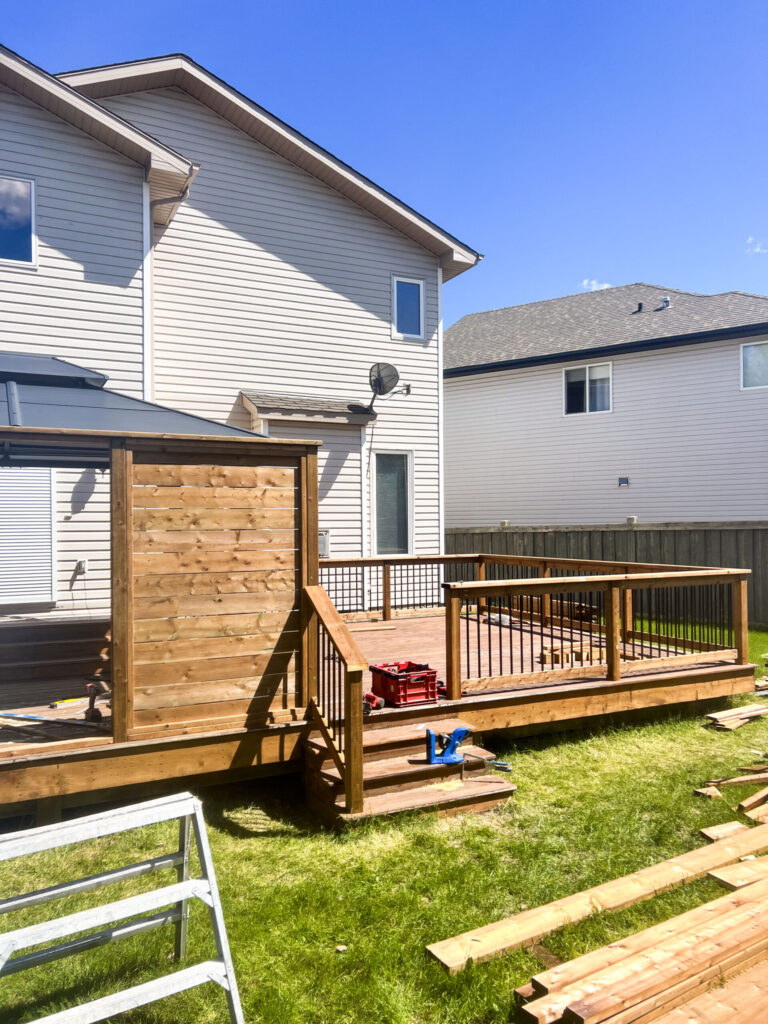 Completed backyard deck in Edmonton with a wooden privacy wall, black aluminum railings, and stairs leading down to the lawn.